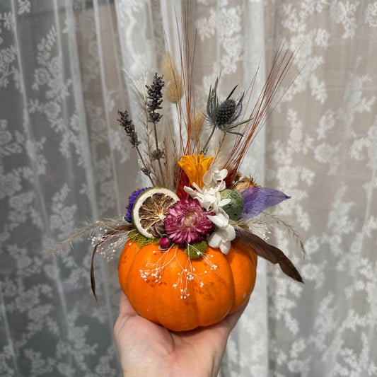 Hand holding a small pumpkin with decorative flowers and feathers against a lace curtain background