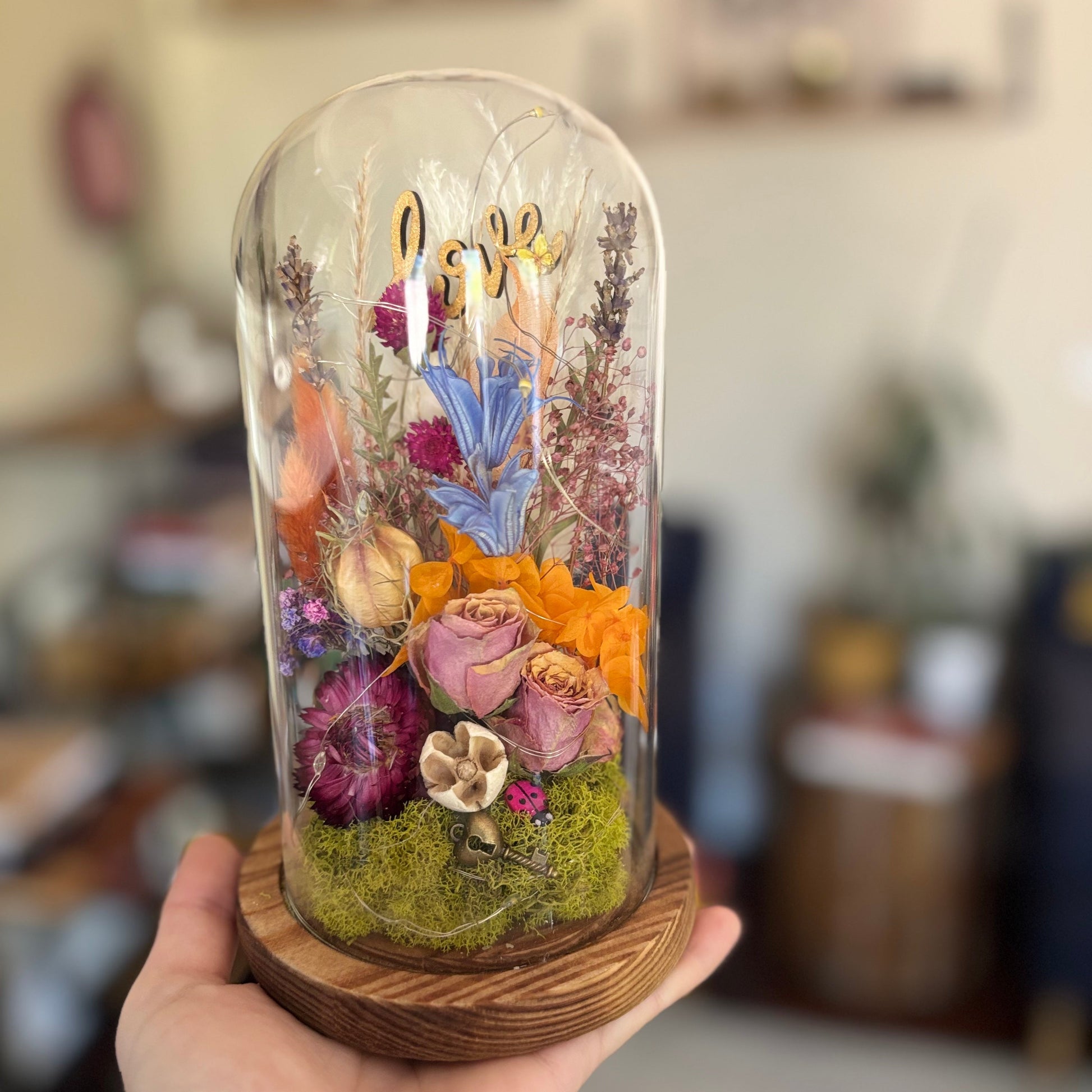 Glass dome with preserved flowers and a butterfly held by a hand in an indoor setting.
