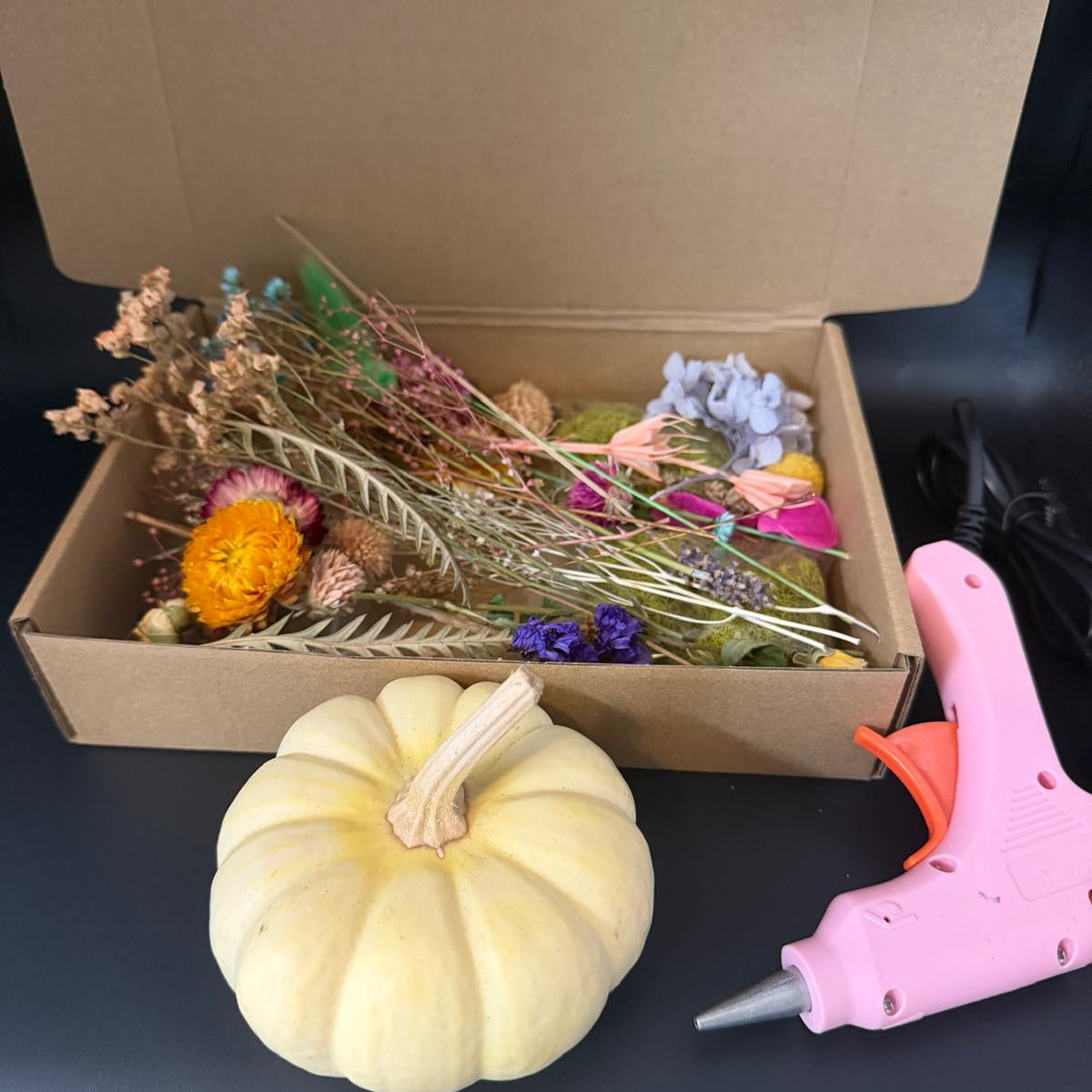 Box of dried flowers, a small pumpkin, and a pink hot glue gun on a dark surface.