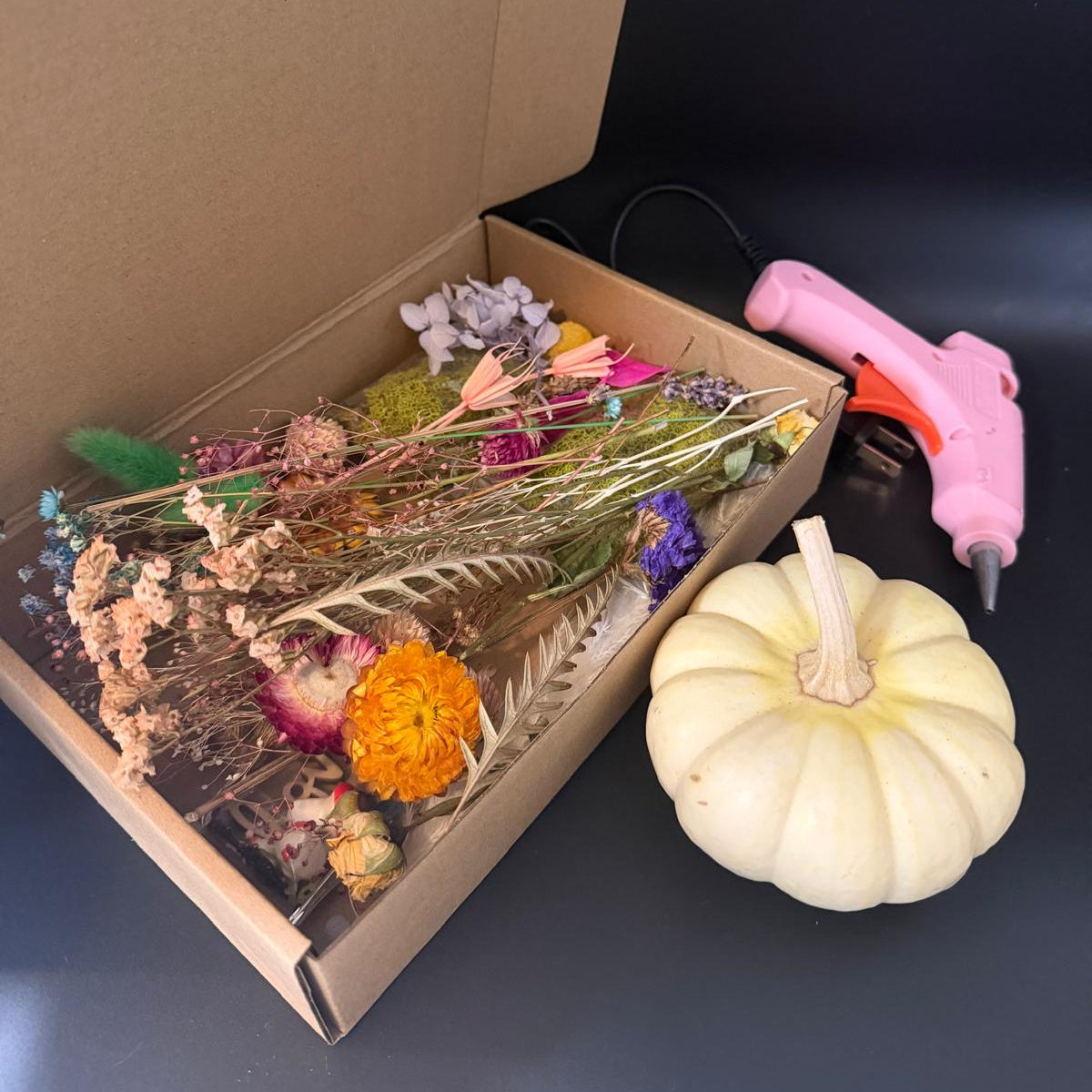 Box of dried flowers, a small white pumpkin, and a pink hot glue gun on a dark surface.